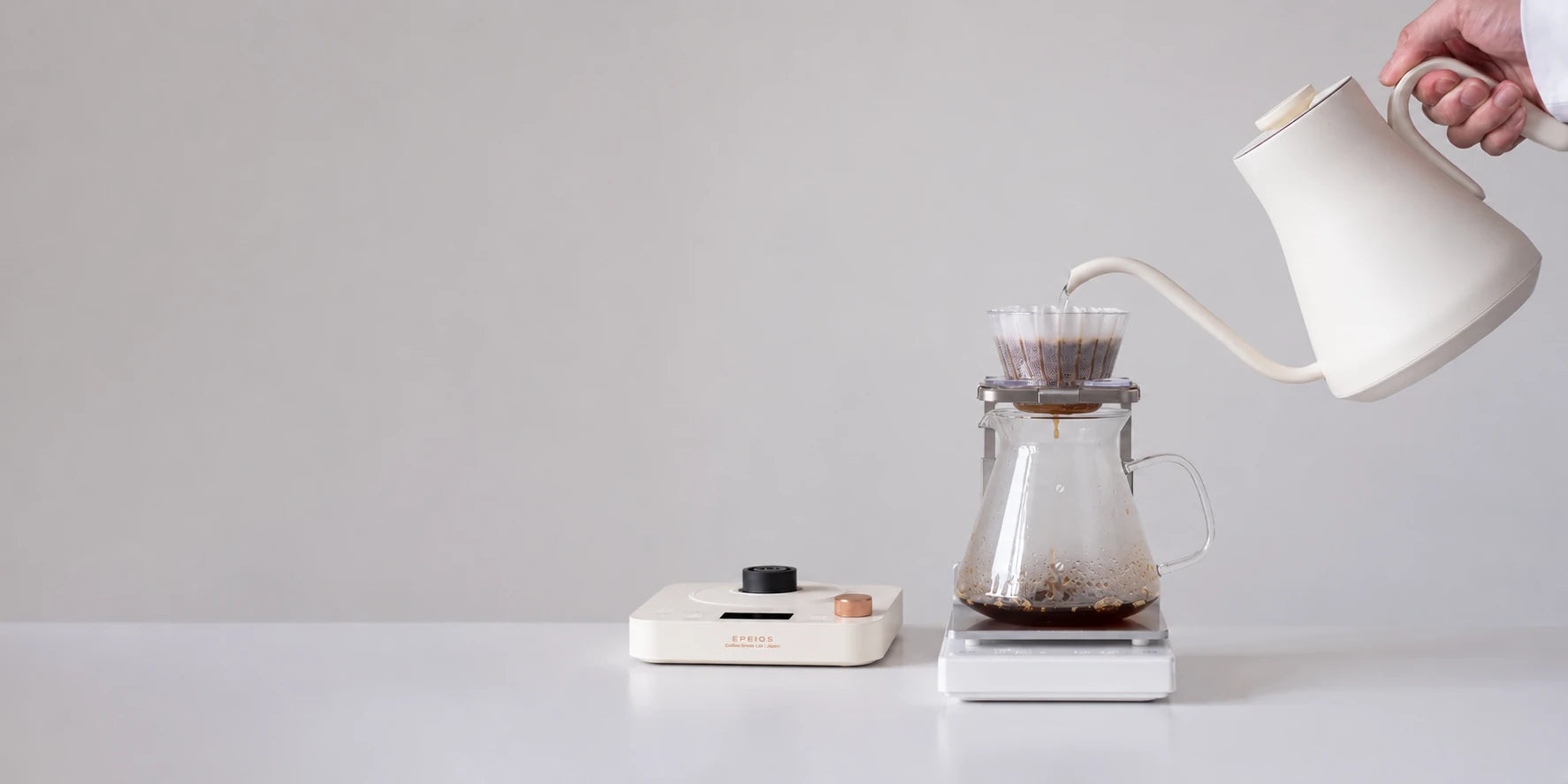 A cream white EPEIOS gooseneck kettle pouring coffee beside its U.S. model control base
